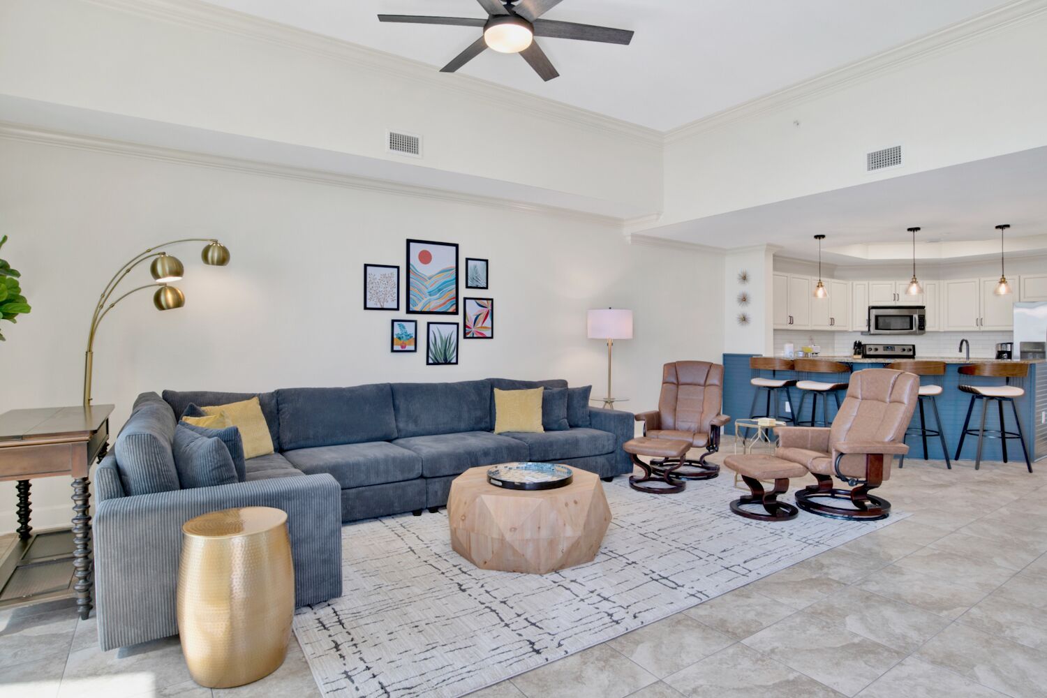 A modern living room with a sofa, coffee table, and chairs, a kitchen with bar stools, and a ceiling fan.
