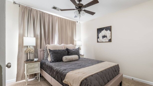 Une chambre à coucher avec un lit, un ventilateur de plafond, une table de chevet, une lampe, un tableau et une porte.