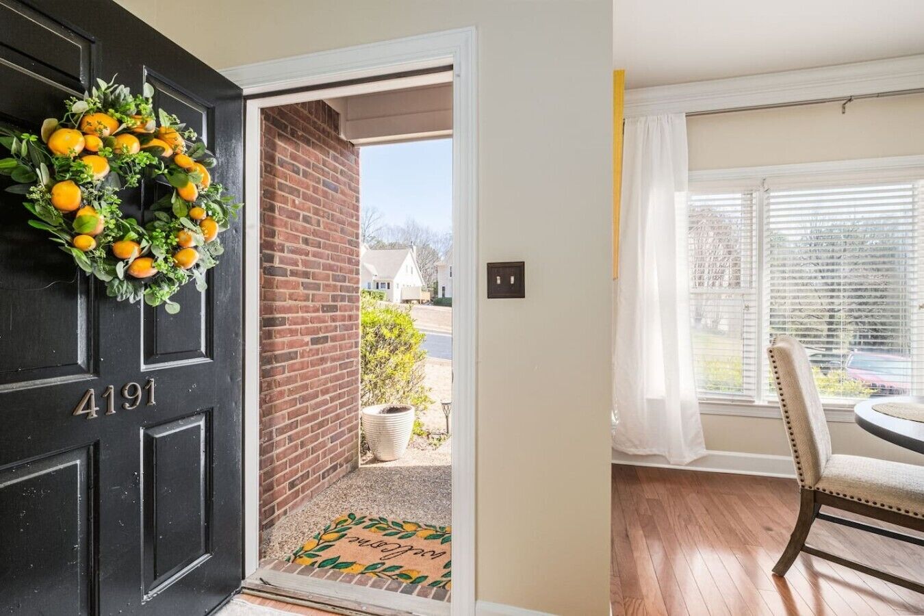 Eine schwarze Tür mit der Nummer 4191, ein Kranz mit Orangen, eine Ziegelwand, eine Fußmatte, ein Fenster mit Rollläden und ein Essbereich mit Stuhl und Tisch.