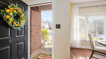 Eine schwarze Tür mit der Nummer 4191, ein Kranz mit Orangen, eine Ziegelwand, eine Fußmatte, ein Fenster mit Rollläden und ein Essbereich mit Stuhl und Tisch.