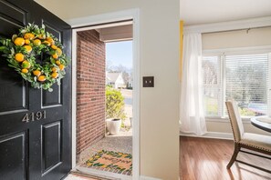 A black door with the number 4191, a wreath with oranges, a brick wall, a doormat, a window with blinds, and a dining area with a chair and table.