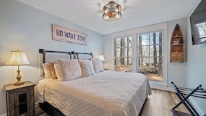 Ein Schlafzimmer mit einem großen Bett, zwei Nachttischlampen, einem Schild mit der Aufschrift "NO WAKE ZONE", einem Fenster mit Blick auf Bäume und einer Holzablage.