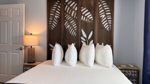 A hotel room with a large bed, a wooden headboard with leaf patterns, white pillows, a bedside lamp, and a nightstand with a remote control.