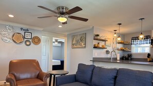 A living room with a brown leather armchair, a blue sofa, and a coffee table. There is a ceiling fan, a painting, and a kitchen area visible in the background.