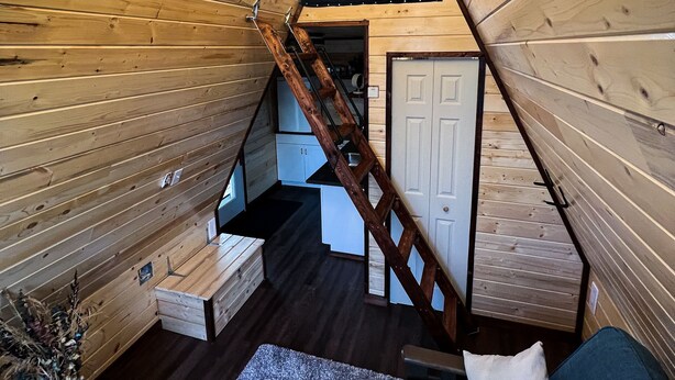 A cozy cabin interior with a loft bedroom, wooden walls, a staircase, a small kitchen, and a seating area with a blue sofa and a white pillow. - Tiny A-Frame Cabin with Outdoor Fire Pit! (Sunset House)