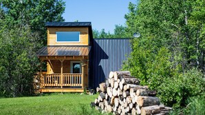 A wooden cabin with a balcony, a small deck, and a stack of firewood in a grassy area with trees. - Tiny A-Frame Cabin with Outdoor Fire Pit! (Sunset House)