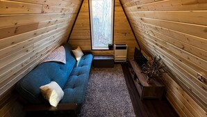 A cozy attic bedroom with a sloped ceiling, a blue sofa, a small bedside table, and a window with a view of trees. - Tiny A-Frame Cabin with Outdoor Fire Pit! (Sunset House)