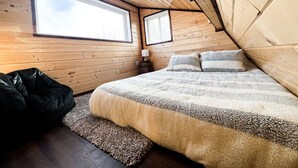 A cozy bedroom with a large window, wooden walls, a bed with fluffy bedding, and a small bedside table. - Tiny A-Frame Cabin with Outdoor Fire Pit! (Sunset House)
