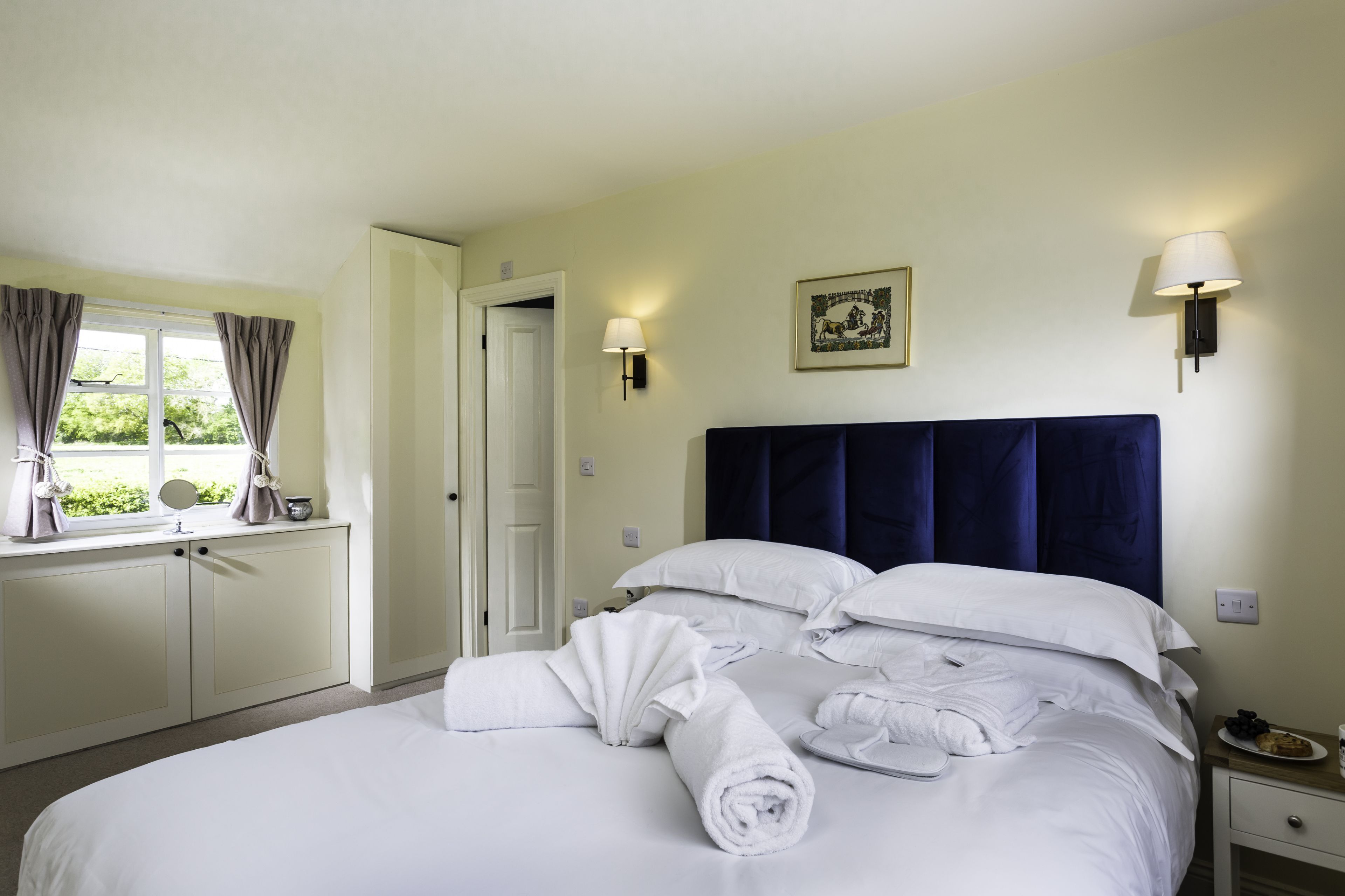 A neatly made bed with white linens and towels, a bedside table with a lamp, and a window with curtains.