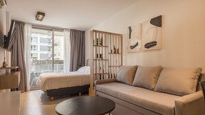 A modern hotel room with a bed, sofa, and a small table. There is a balcony with a view of buildings. - CloudBA Guemes Soho Apartments (Buenos Aires)