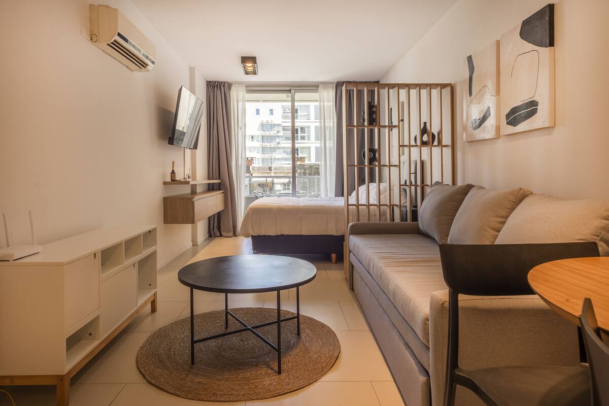 A modern, well-lit bedroom with a bed, a sofa, a small table, and a TV.