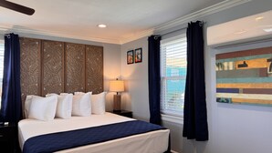 Un dormitorio con cama, mesita de noche, lámpara, panel decorativo, ventana con cortinas y una obra de arte colgada en la pared.