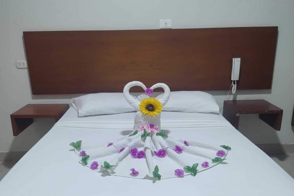 A neatly made bed with a towel arranged in a heart shape and a sunflower centerpiece, a wooden headboard, and a small bedside table with a phone.
