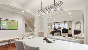 Ein geräumiges Wohnzimmer mit großem weißen Tisch, bequemen Sitzgelegenheiten, einem Klavier, einem Fernseher und einem Kamin.