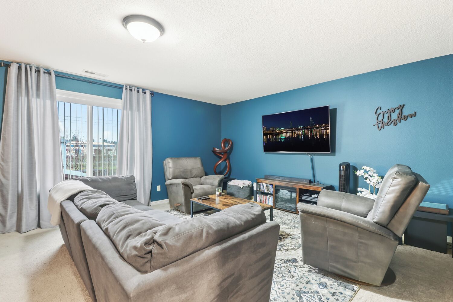 Un salon avec un mur bleu, une grande fenêtre, un canapé, deux fauteuils, une table basse et un téléviseur à écran plat.