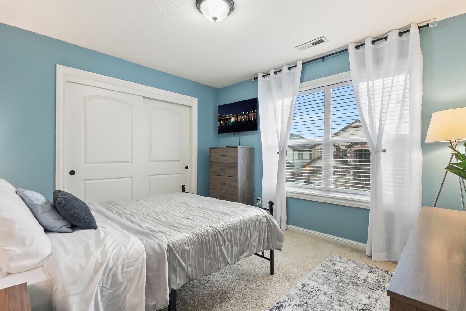 A bedroom with a bed, a dresser, a TV on the wall, and a window with curtains.