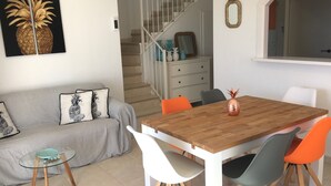 Un salon avec une table à manger en bois, des chaises blanches et orange, un canapé gris agrémenté de coussins à motifs d’ananas, et un escalier en arrière-plan.