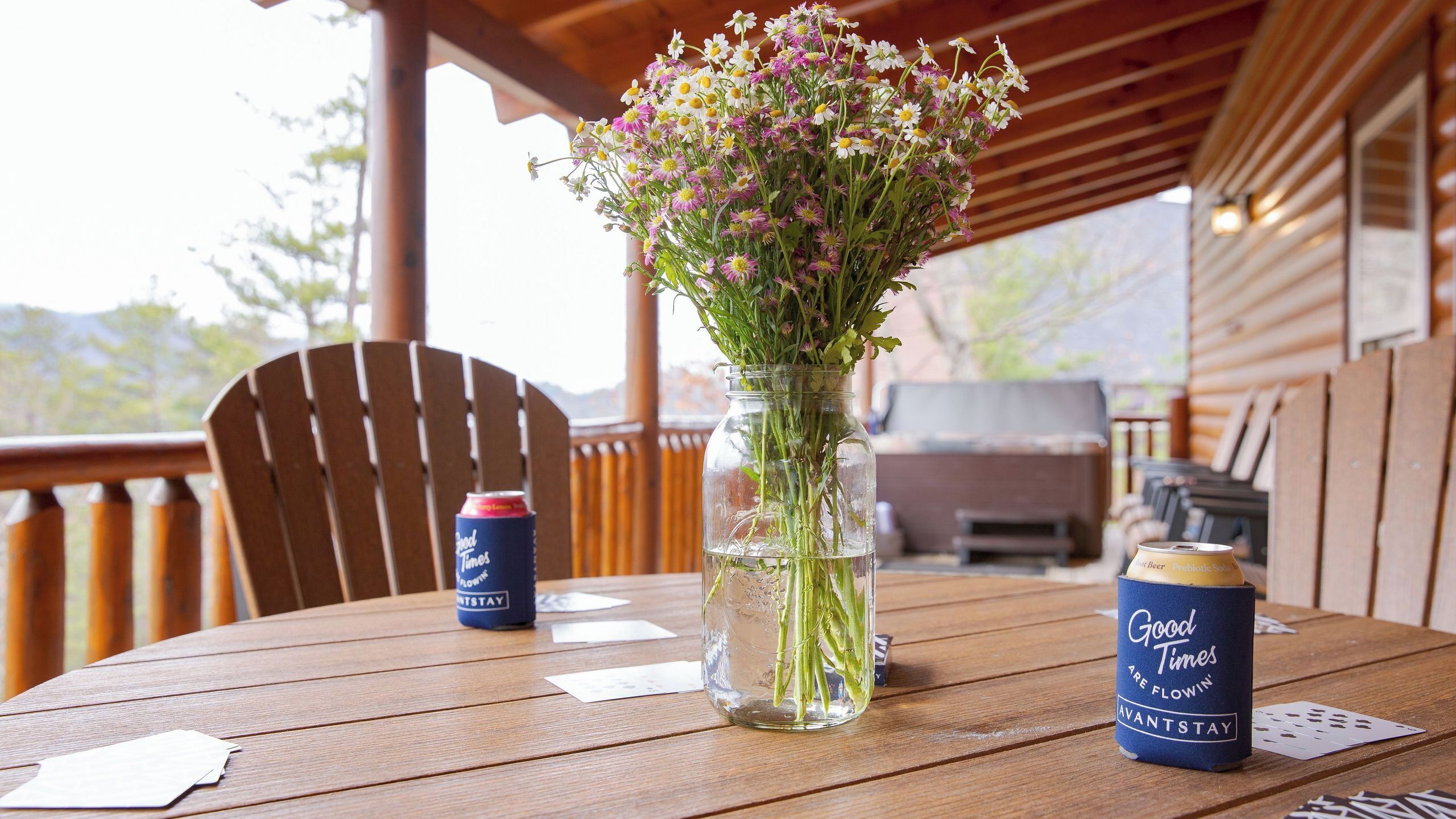 A wooden table with a glass vase of flowers, two cans of beer, and playing cards.