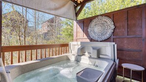 Een hottub met een stenen rand, een klein tafeltje en een decoratieve wandplaat, gelegen op een houten terras met uitzicht op bomen en gebouwen.