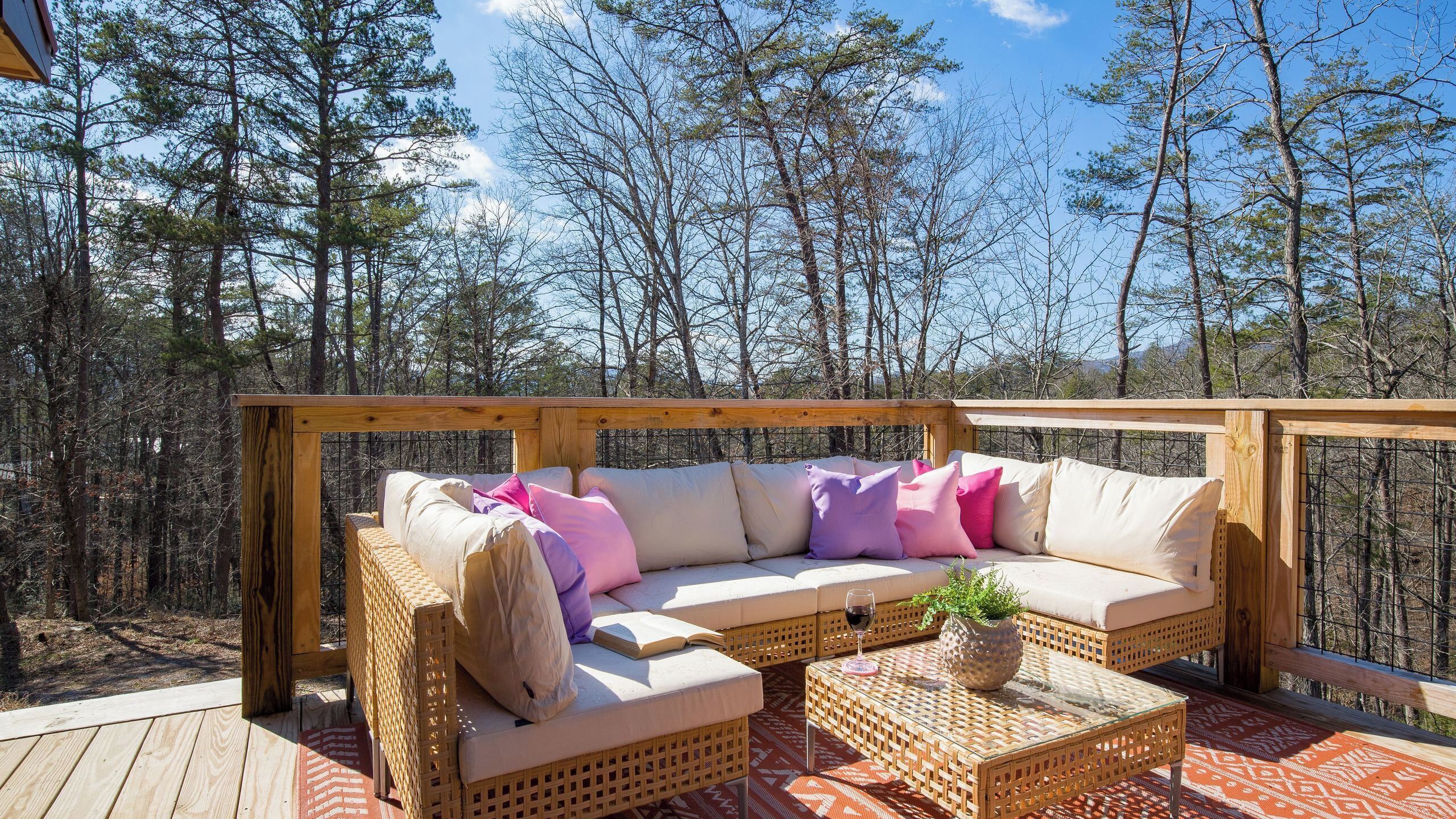 A wooden deck with a wicker sofa and a glass coffee table, surrounded by a forest.