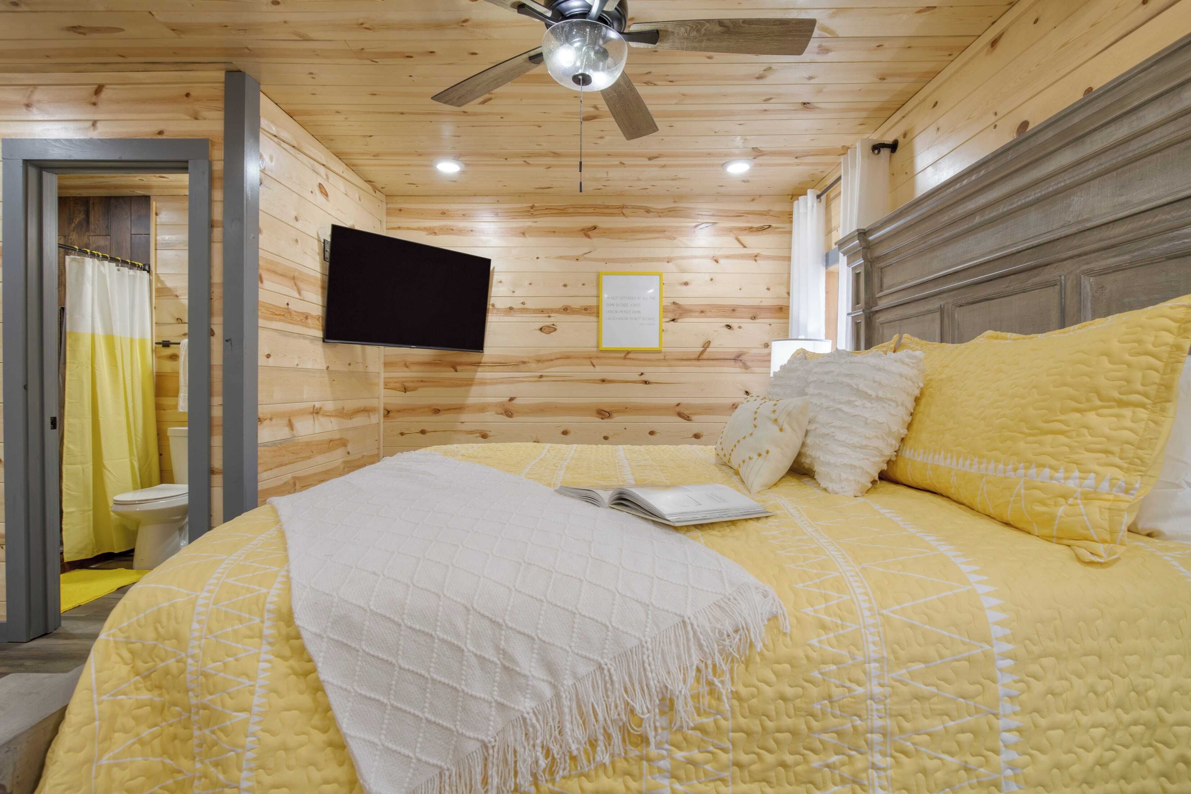 Une pièce aux murs lambrissés, comprenant un lit, un ventilateur de plafond, un téléviseur à écran plat et un rideau de douche à rayures jaunes et blanches.