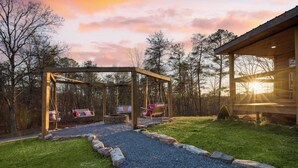 A wooden gazebo with swings, a fire pit, and a gravel path leading to a cabin. - Pink Pines by Avantstay Luxury Aesthetic Dream Home, Indoor Pool, Hot Tub (Sevierville)