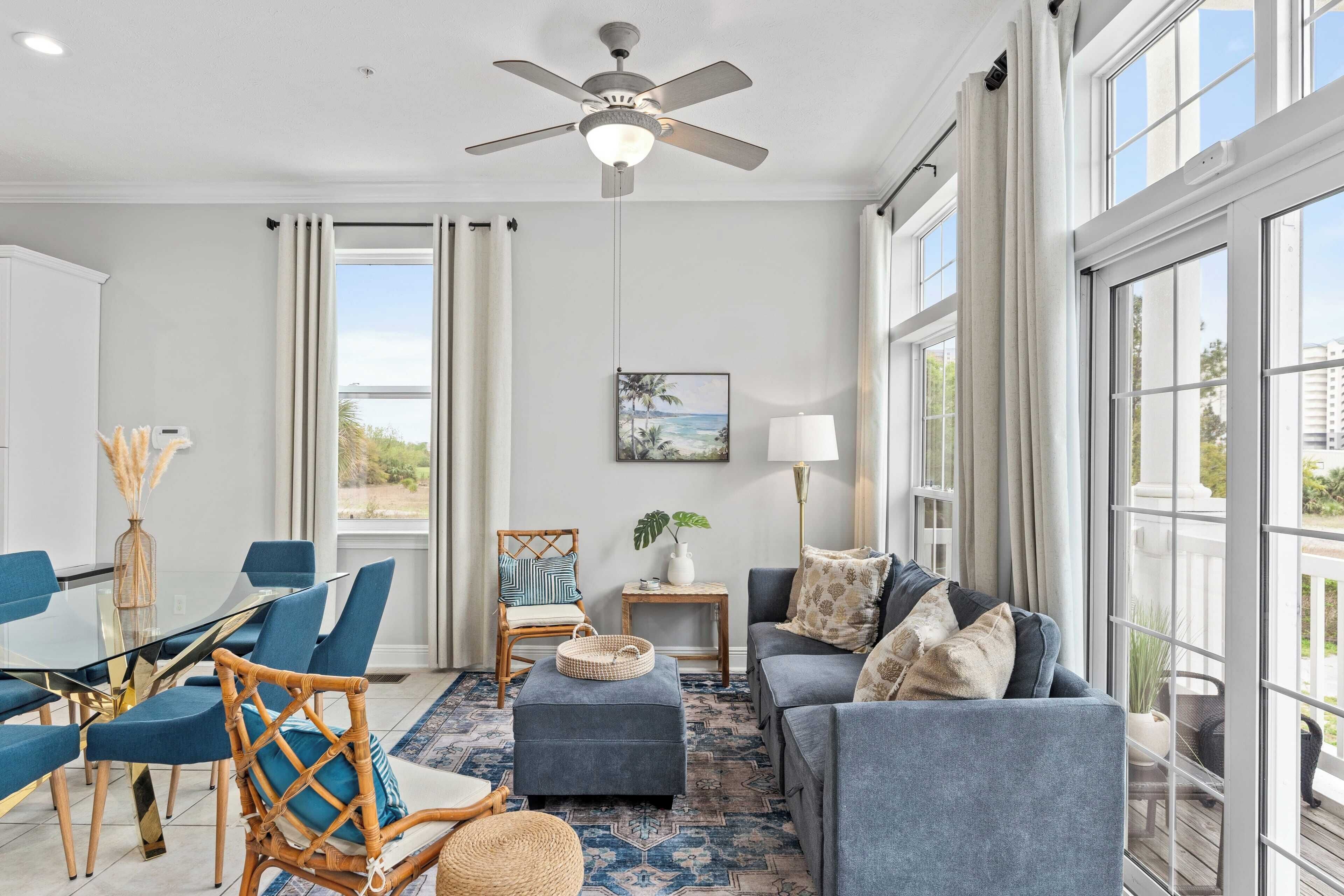 Un vaste salon doté d’un ventilateur de plafond moderne, de grandes fenêtres, d’un coin repas avec des chaises bleues et d’un espace salon confortable avec un canapé bleu et un pouf.