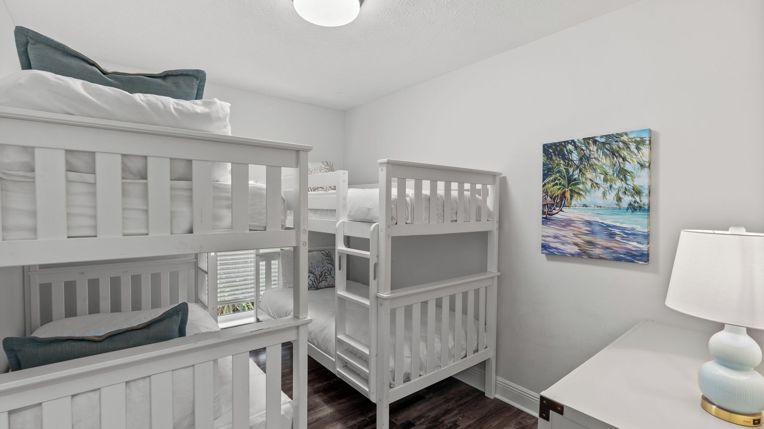 Une chambre avec deux lits superposés blancs, un bureau blanc, une lampe et un tableau sur le thème de la plage accroché au mur.