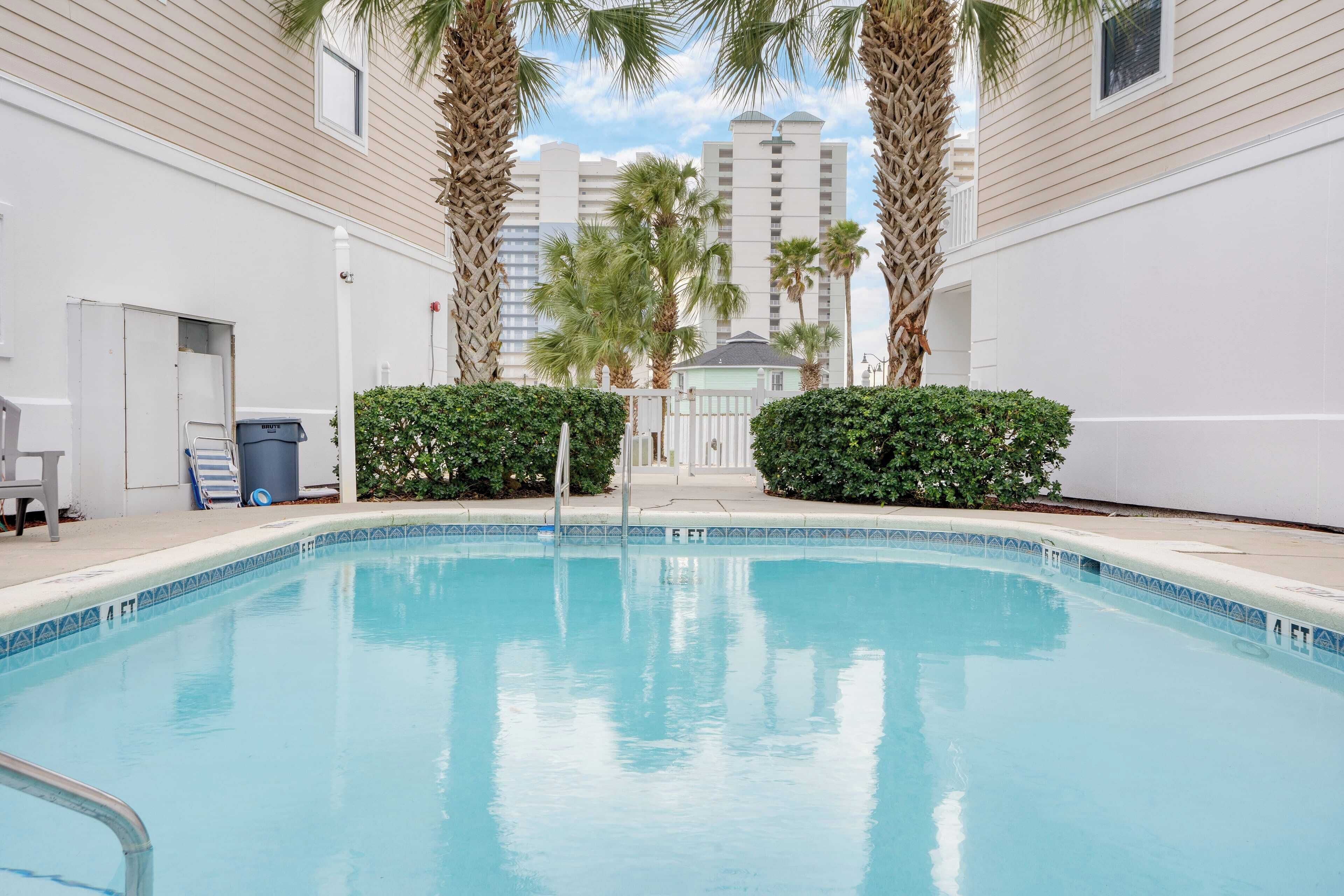 Une piscine dont le bord est recouvert de carrelage, entourée de palmiers et d’une clôture blanche.