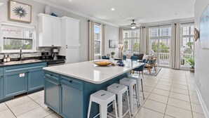 A modern kitchen with blue cabinets, a large island, and a dining area with a view of the city. - Tidal Charm by Avantstay Walk to Beach, Community Pool, Balcony, Ocean Views (Panama City Beach)