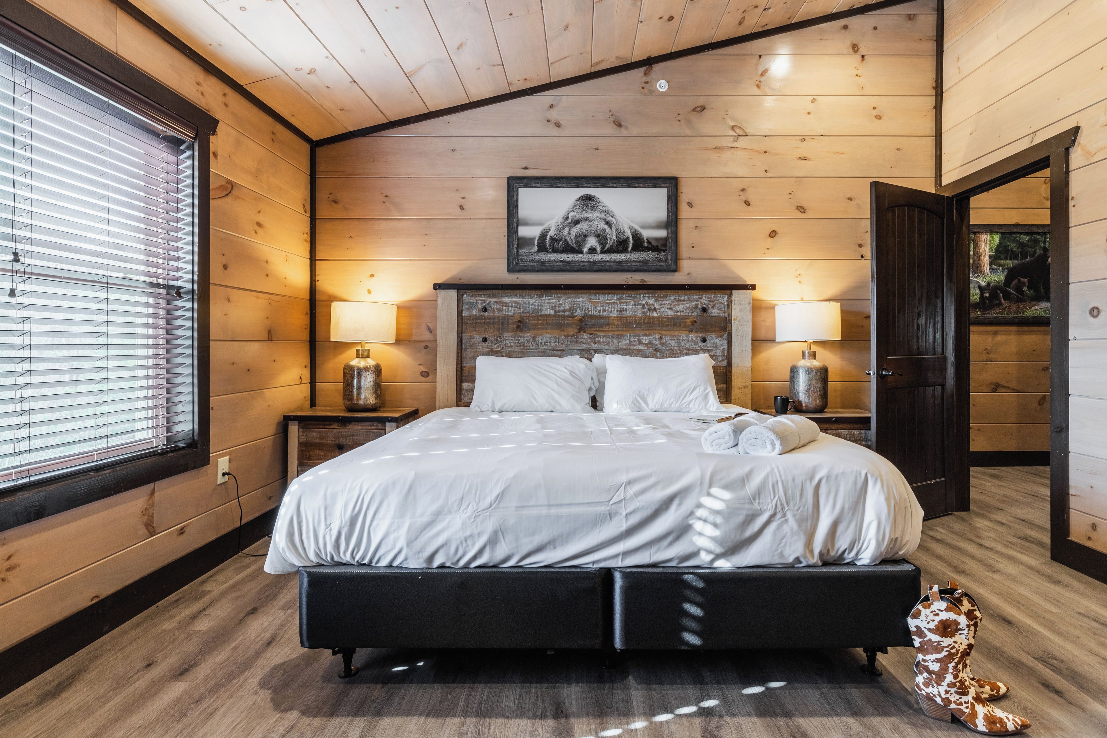 A wooden-paneled bedroom with a bed, two bedside lamps, a framed picture above the bed, and a window with blinds.
