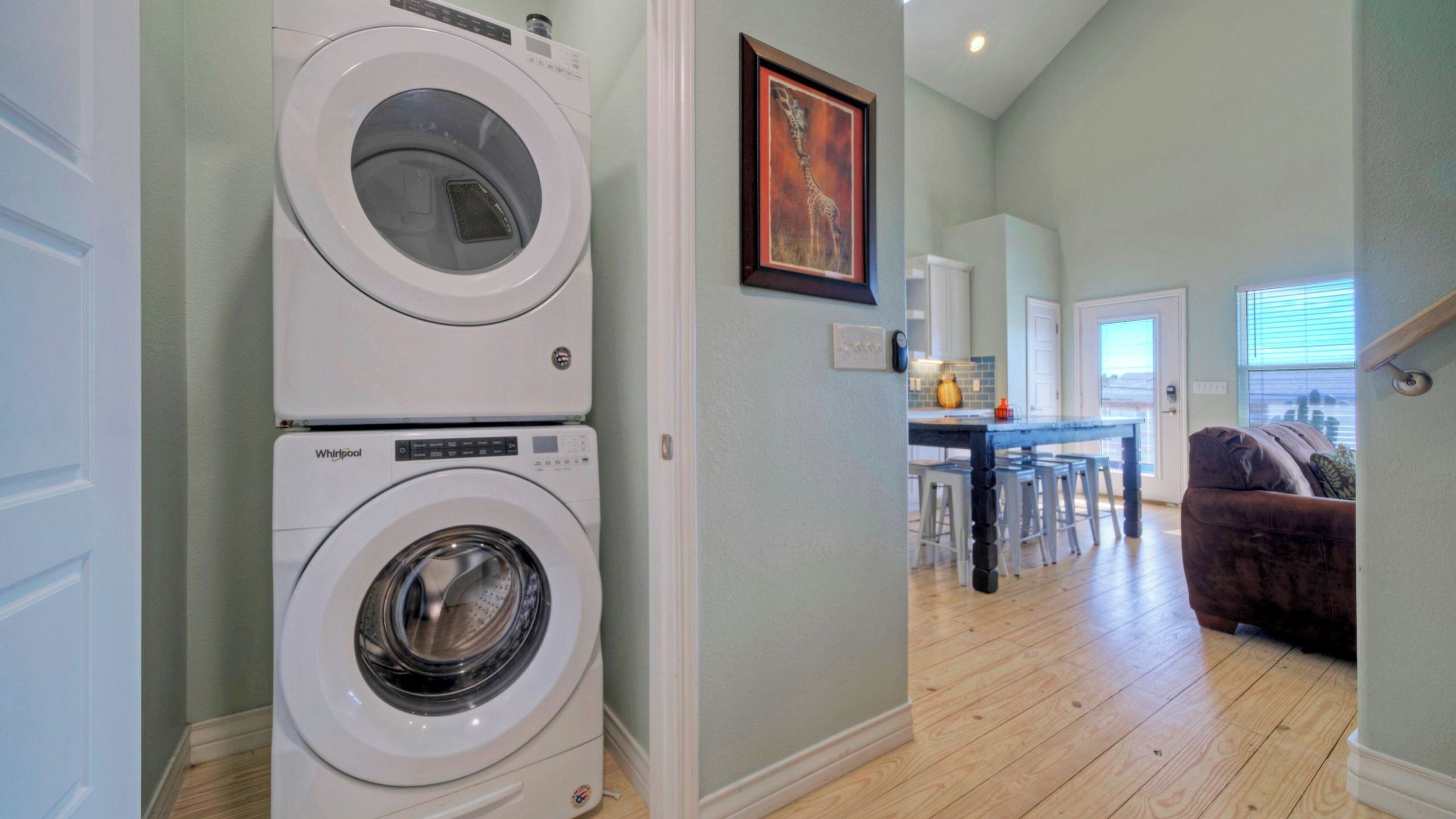 A laundry room with two stacked Whirlpool washers, a dining area with a table and chairs, and a living room with a brown sofa.