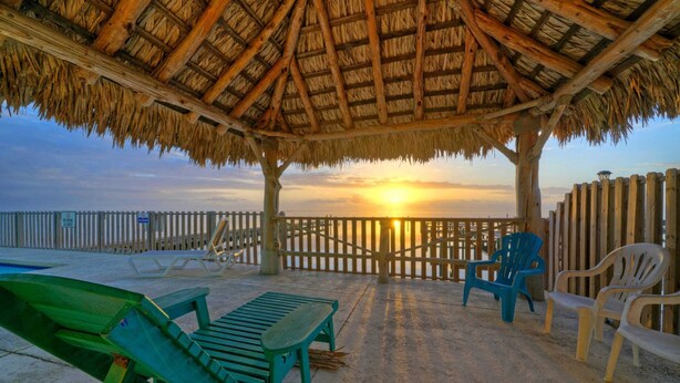 Un patio frente a la playa con techo de paja, sillones de descanso y vista al atardecer sobre el océano.