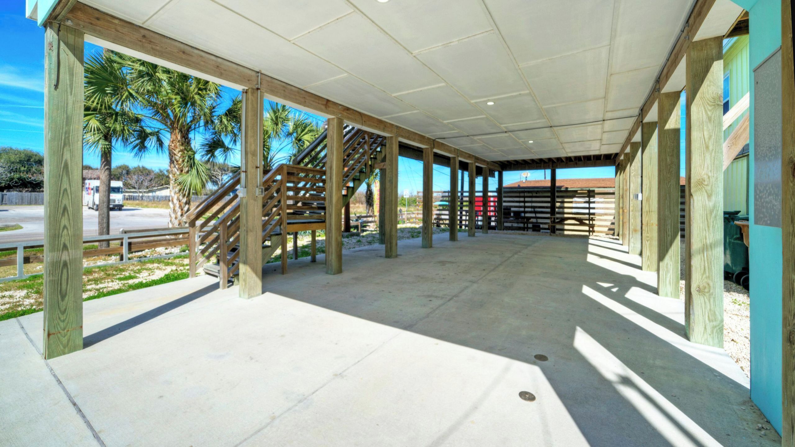 Un espacio al aire libre techado con columnas de madera, piso de concreto y vista a palmeras.