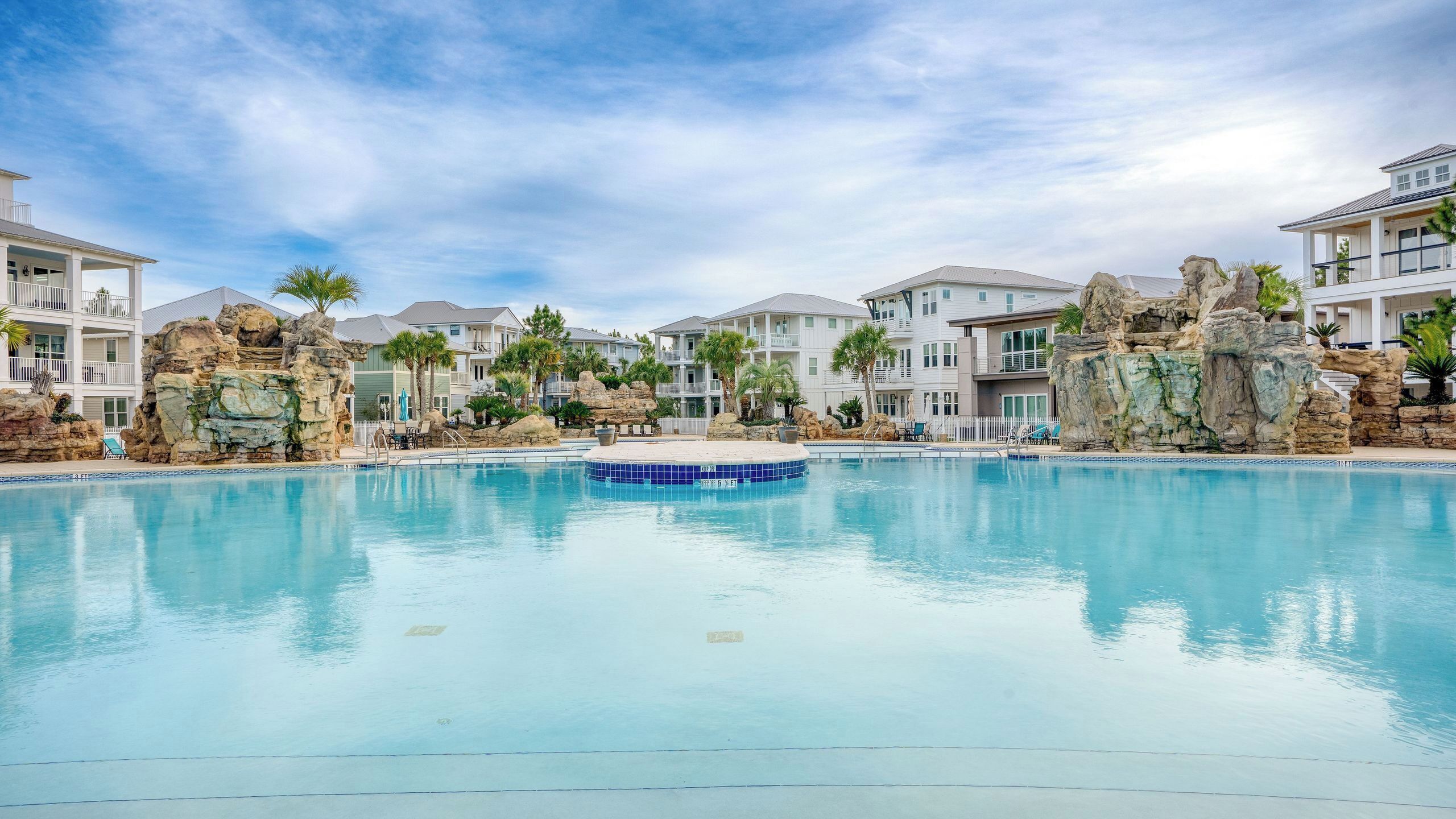 A swimming pool with a central island, surrounded by buildings with balconies and rock formations.