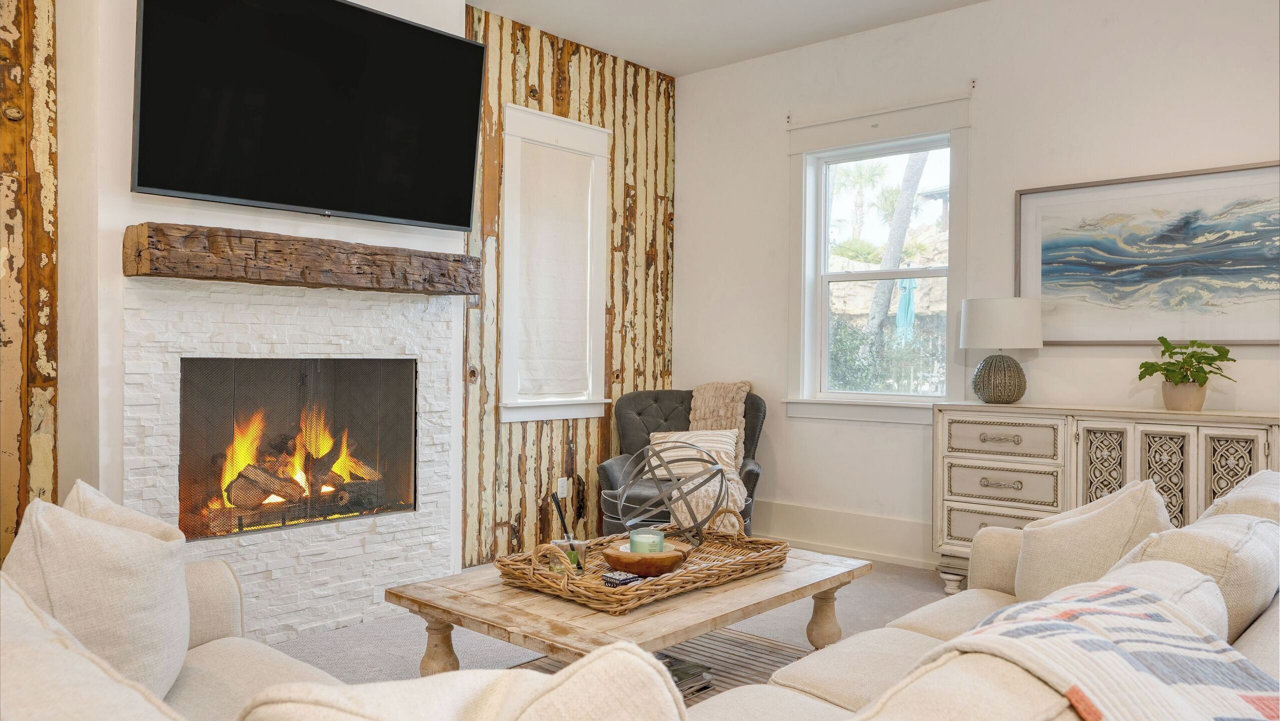 A living room with a fireplace, a television mounted above it, a sofa set, a coffee table with decorative items, and a window with a view of trees.