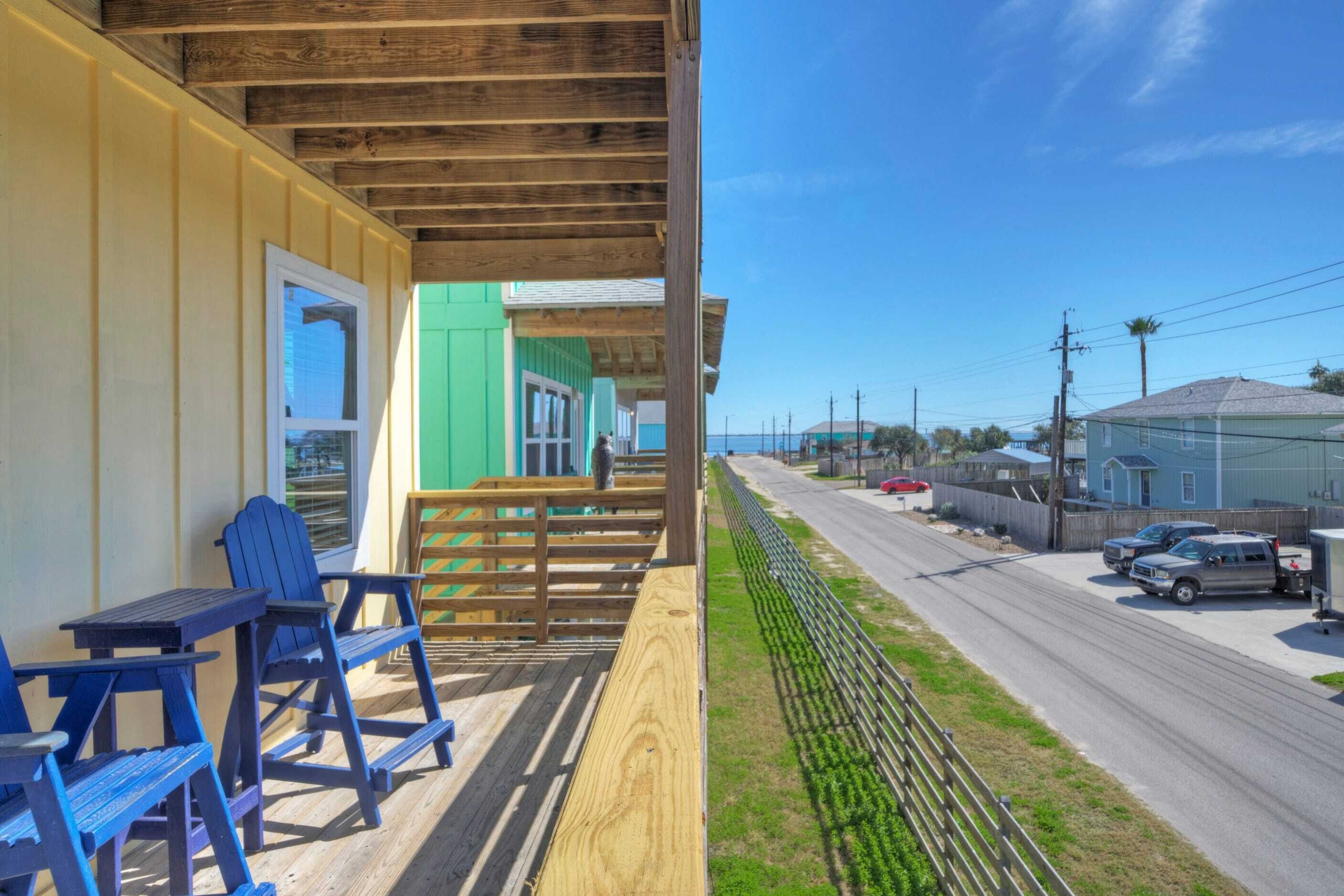 A wooden deck with blue Adirondack chairs, a wooden bench, and a view of a street with houses and parked cars.