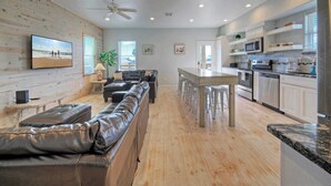 A modern kitchen with white cabinets, a dining area with a wooden table, and a living room with a leather sofa. - 6bdr, Balcony w/ Bay View, Shared Pool + Boat Dock Keeper Red by Avantstay (Corpus Christi)