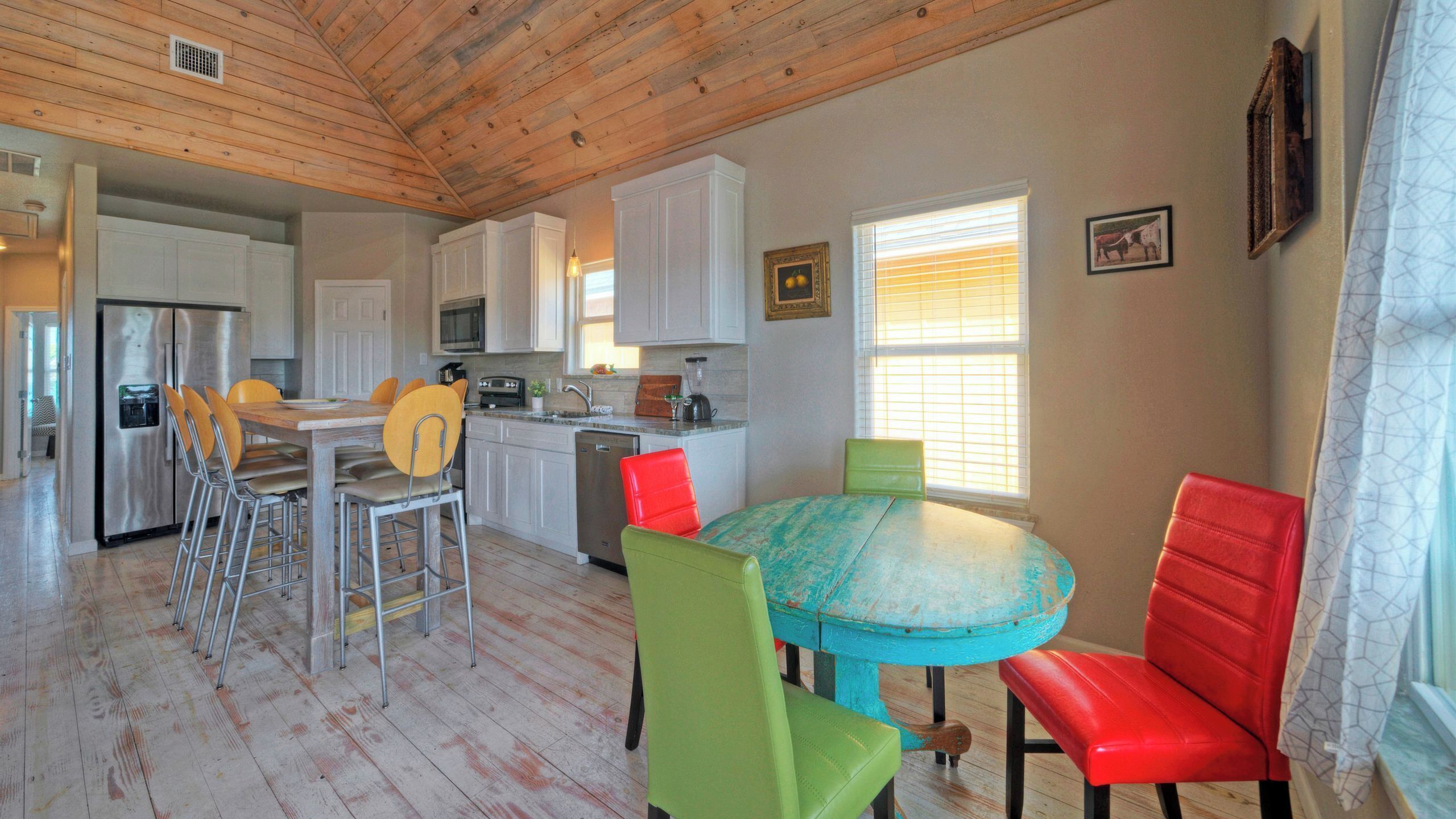 A kitchen with white cabinets, a stainless steel refrigerator, a sink, and a dining area with a round table and colorful chairs.