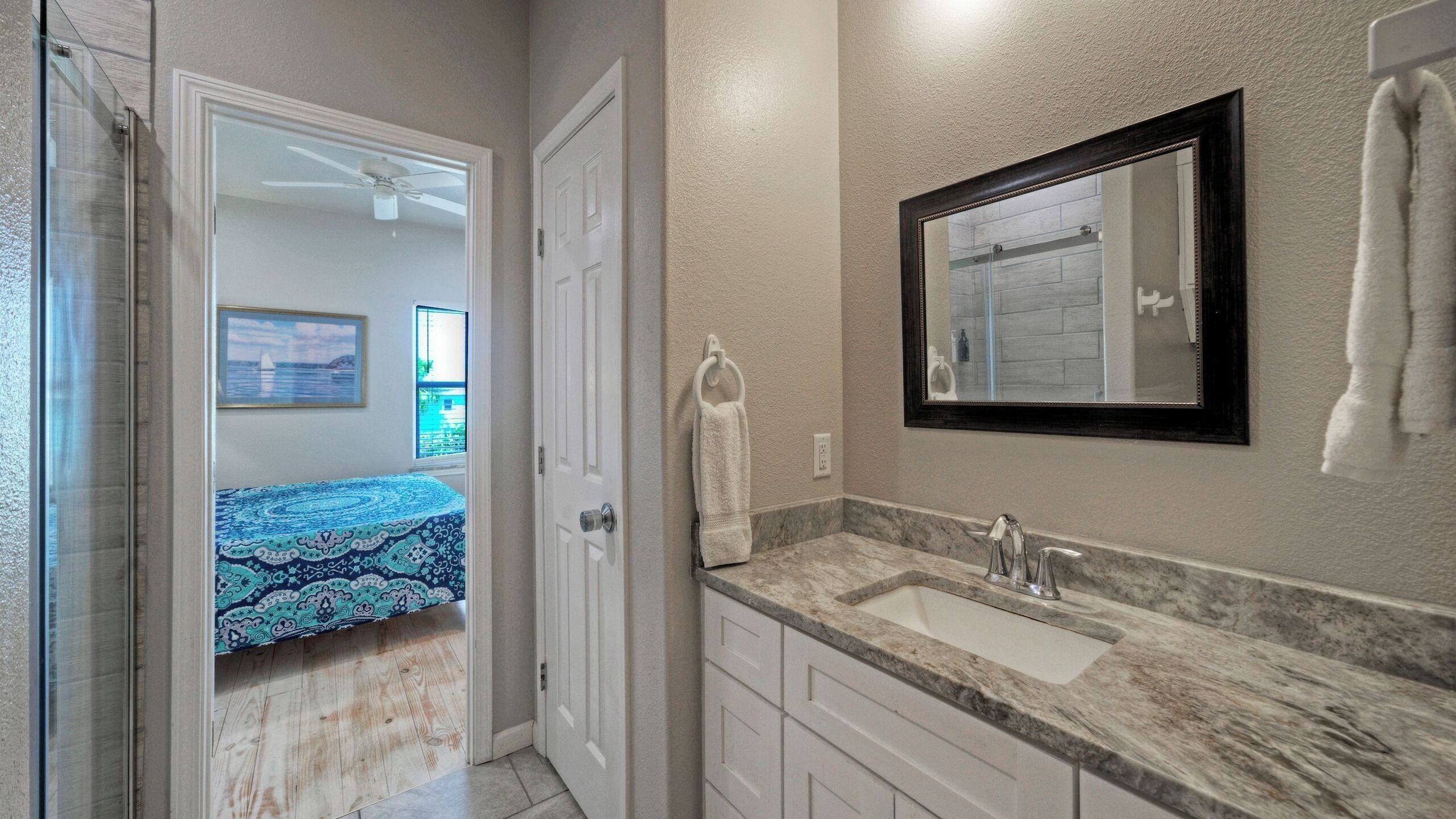 A bathroom with a sink, mirror, towel rack, and a door leading to a bedroom with a bed and a window.