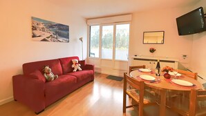 Una sala de estar con un sofá rojo, un osito de peluche, un cuadro, una mesa de comedor preparada para cuatro personas y un televisor montado en la pared.