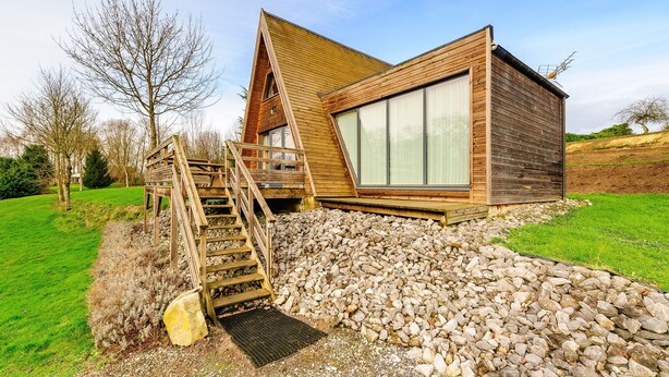 Una cabaña moderna de madera con techo inclinado, un ventanal grande y una escalera de madera que conduce a una terraza.
