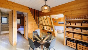 Interior moderno de cabaña de troncos con mesa de comedor de cristal, estanterías de madera y un dormitorio en el altillo.
