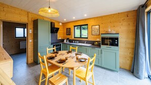 Un interior de madera con una mesa redonda de comedor, sillas, una zona de cocina con armarios verdes y una lámpara colgante.