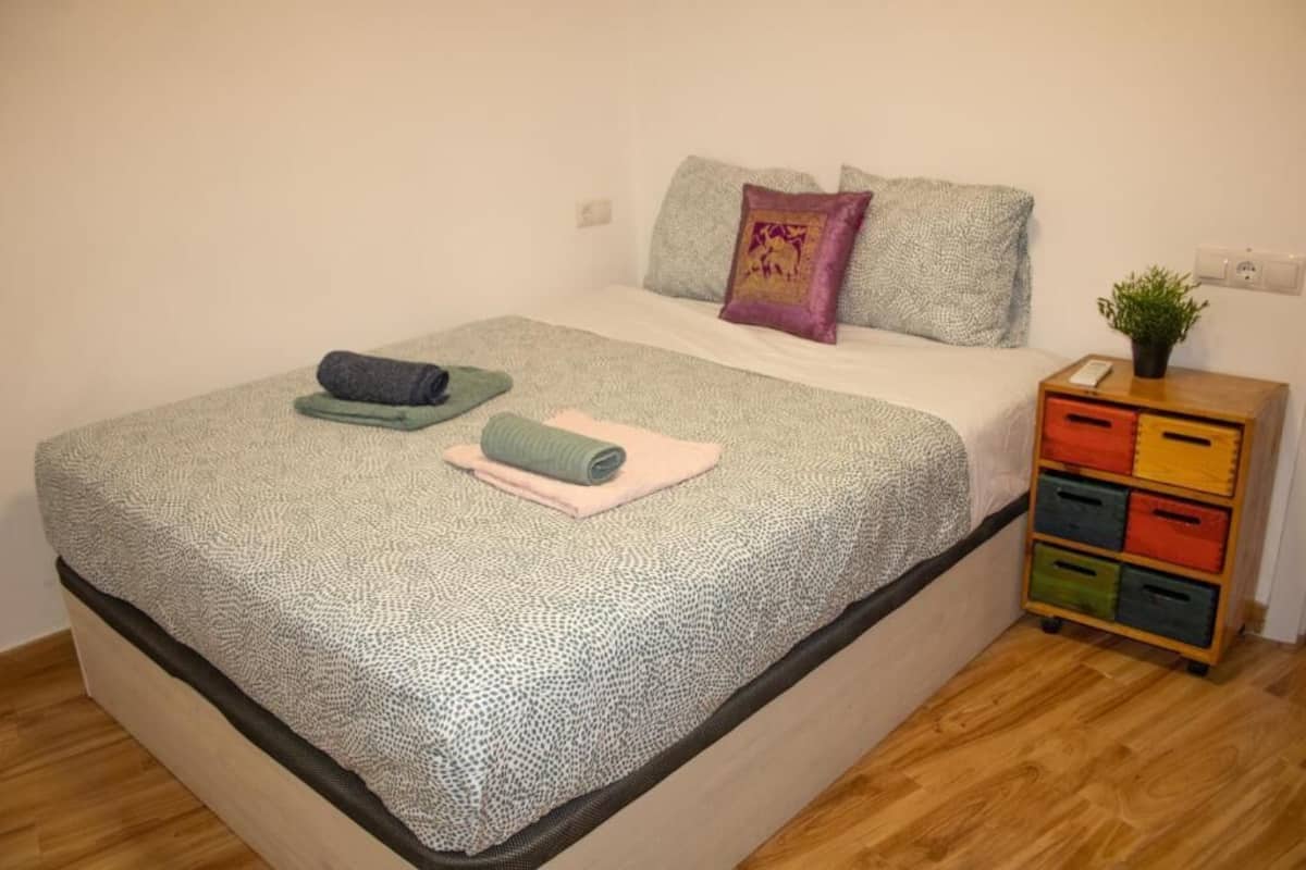Un lit bien fait, recouvert d’une couvre-lit à motifs, une petite table de chevet avec une plante et un meuble de rangement coloré.
