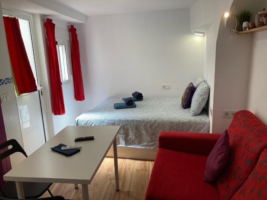 Ein kleines Zimmer mit einem Bett, einem kleinen Tisch, einem roten Sofa und einem Fenster mit roten Vorhängen.