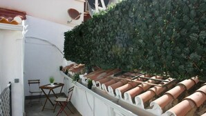 Una terraza en la azotea con baldosas de terracota, una pared blanca con seto frondoso y una pequeña mesa con sillas.