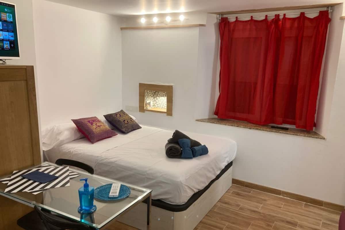 Une petite chambre comprenant un lit, une petite table, un téléviseur et une fenêtre avec des rideaux rouges.