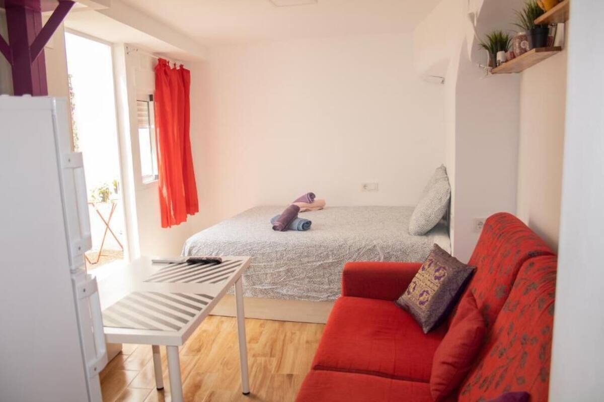 Un espace de vie compact comprenant un lit, un canapé rouge, une petite table et une fenêtre avec des rideaux.