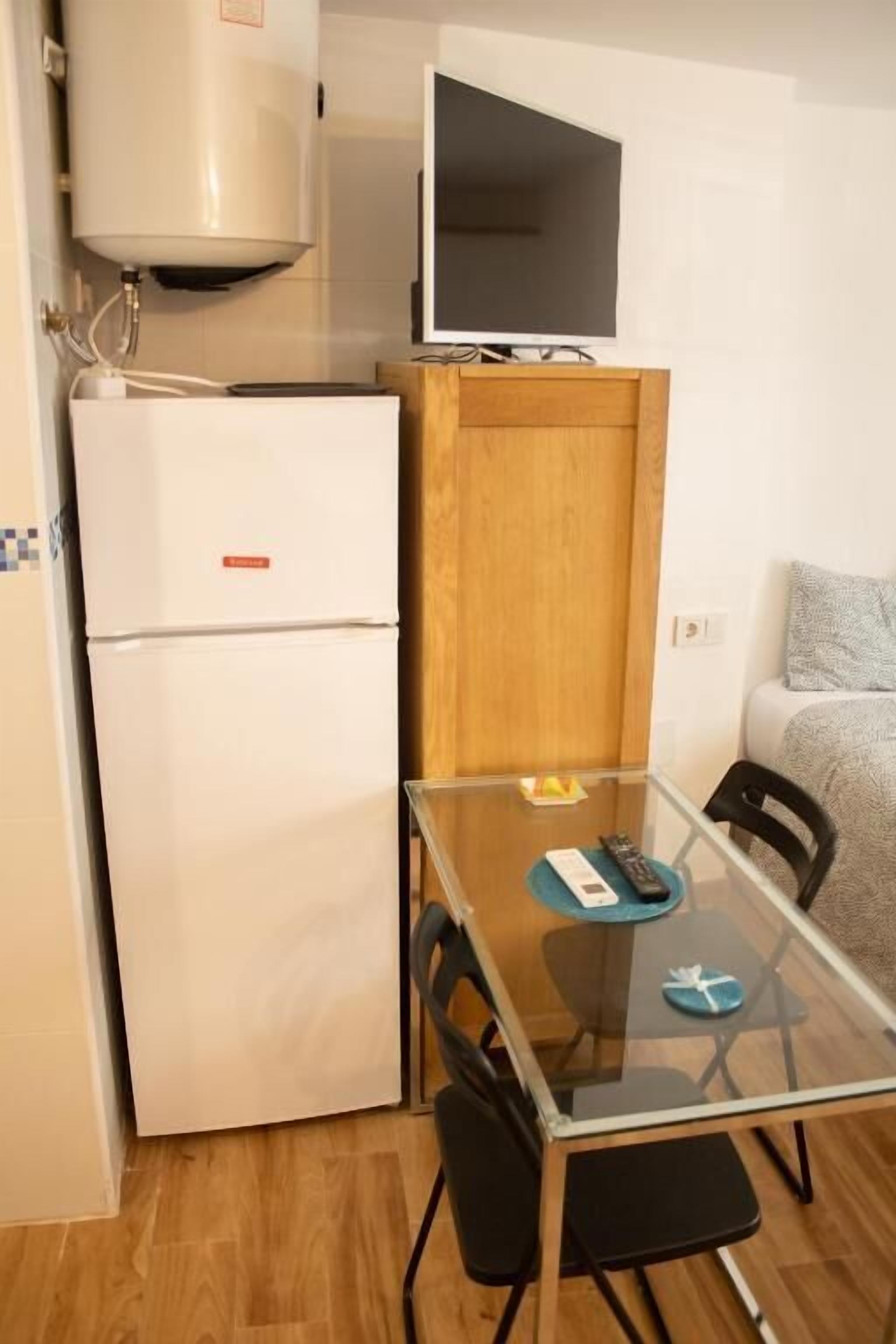 Una cocina compacta con un refrigerador blanco, un mueble de madera, una mesa de vidrio con sillas y un televisor fijado a la pared.